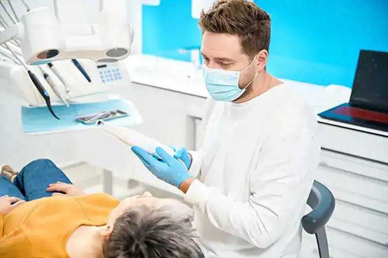 Male dentist wearing a face mask and gloves, using an intraoral scanner to capture digital impressions of a patient's teeth in a bright, modern dental office.